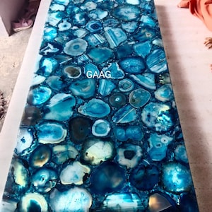 Puede incluir: Un tablero de mesa rectangular hecho de rodajas de ágata azul pulida con un patrón de mosaico. La superficie presenta varios tonos de azul, turquesa y blanco con texturas cristalinas naturales. La palabra GAAG está impresa en blanco.