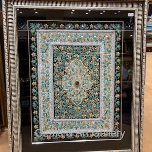 Puede incluir: Una obra de arte enmarcada con un marco adornado en plata y oro. La obra de arte es un diseño bordado en blanco y negro con acentos turquesa. El diseño presenta un motivo floral central con un patrón repetitivo de flores y hojas.