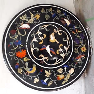 Può includere: Un piano da tavolo rotondo nero con un intarsio decorativo di uccelli, fiori e foglie colorati. L'intarsio è realizzato con diversi tipi di pietra e crea un bellissimo motivo a mosaico.