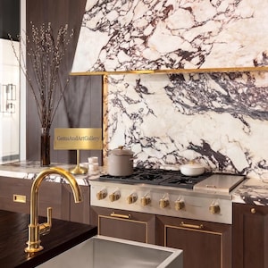 May include: A kitchen scene featuring a stainless steel gas range with gold knobs, a white and purple marble backsplash, dark wood cabinetry with gold hardware, a gold tap, a stainless steel sink, and a gold sign reading GemsAndArtGallery.