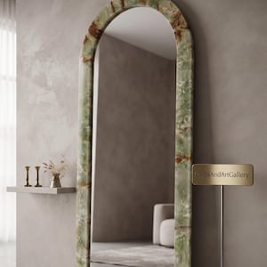 May include: A tall, arched floor mirror featuring a frame made of polished green onyx with natural brown and white veining. A small gold-toned sign stands to the right of the mirror, clearly displaying the text GemsAndArtGallery in black font.