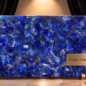 May include: A large rectangular slab of deep blue lapis lazuli stone with visible gold-colored pyrite inclusions. The slab is supported by two small gold-toned metal stands. A gold-colored sign stands in front with the text GemsAndArtGallery.