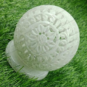 May include: White marble sphere with intricate carved floral designs. The sphere is on a white marble base.