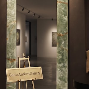 May include: A tall rectangular mirror with a polished light green onyx frame featuring natural brown veining. In front of the mirror stands a gold-toned metal easel holding a rectangular sign with the text GemsAndArtGallery in black font.