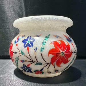 May include: White marble vase with floral inlay. The vase features a wide mouth and a rounded body. The floral design includes red, blue, and green accents. The text "Gems & Art Gallery" is visible on the top of the vase.