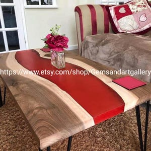 May include: A handcrafted wooden coffee table with a vibrant red epoxy resin river. The table features a natural wood grain and black hairpin legs. A small glass vase with flowers and a red coaster are on the table.