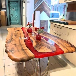 Op de afbeelding: Een houten tafel met een rode epoxyharsrivier die door het midden loopt. De tafel heeft een gouden metalen onderstel en is versierd met rode bloemen in vazen.
