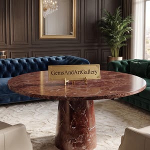 Könnte beinhalten: Ein runder Esstisch aus dunkelrotem Marmor mit einem dicken, zylindrischen Säulenfuß mit weißen Adern. Ein rechteckiges, goldfarbenes Schild mit dem schwarzen Text GemsAndArtGallery steht auf einem kleinen goldenen Ständer in der Tischmitte.