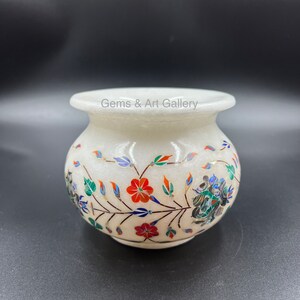 May include: White marble vase with intricate floral designs inlaid with colorful gemstones. The vase has a round body and a wide opening. "Gems & Art Gallery" is written on the vase.