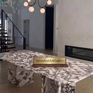 Puede incluir: Una mesa de comedor rectangular con una superficie blanca con vetas de color púrpura oscuro y patas a juego tipo losa. Una placa rectangular de tono dorado con el texto GemsAndArtGallery se asienta sobre una base de madera oscura.