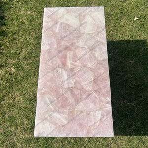 May include: A pink and white rose quartz table top with a natural, irregular, and textured surface. The table top is made of many pieces of rose quartz that have been glued together.