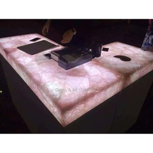 May include: A large, rectangular countertop made of pink quartz with a white, glowing light underneath. The countertop is lit from below, highlighting the natural patterns and textures of the quartz. The countertop is perfect for a retail store or a home bar.