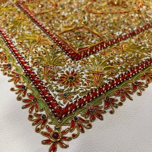 Puede incluir: Un fondo de cuero blanco con un dise&ntilde;o bordado en rojo, dorado y verde. El dise&ntilde;o presenta intrincados patrones florales y peque&ntilde;as piedras preciosas.
