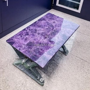 Può includere: Un tavolino rettangolare in ametista con superficie lucida. Il tavolo ha una vibrante tonalità viola con venature bianche. La base è di colore grigio scuro. Il testo "Gems & Art Gallery" è visibile sul tavolo.