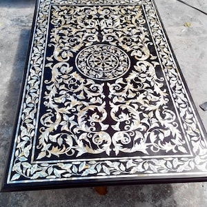 May include: A black table top with a decorative inlay of white and gold mother of pearl. The inlay features a floral design with a central circular motif.