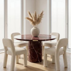 May include: A round dining table with a red marble top and a matching pedestal base. Four cream-colored, curved-back chairs surround the table. A white vase with dried pampas grass sits in the center of the table. The room has large windows.