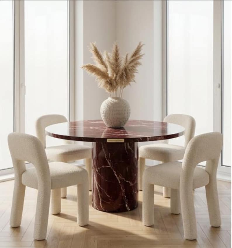 Peut inclure: Une table &agrave; manger ronde avec un plateau en marbre rouge et un piedestal assorti. Quatre chaises cr&egrave;me &agrave; dossier incurv&eacute; entourent la table. Un vase blanc avec des herbes de la pampa s&eacute;ch&eacute;es est au centre. La pi&egrave;ce a de grandes fen&ecirc;tres.