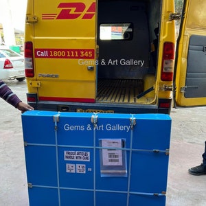 Op de afbeelding: Een blauwe kartonnen doos met de tekst "Gems & Art Gallery" en "Fragile Articles Handle With Care" erop gedrukt. De doos is vastgemaakt met witte tape en wordt in een gele bestelwagen geladen.