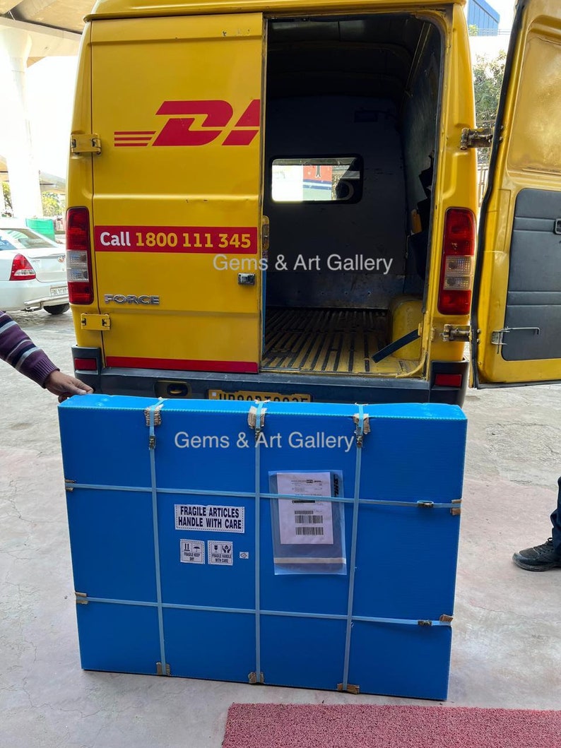 Op de afbeelding: Een blauwe kartonnen doos met de tekst "Gems & Art Gallery" en "Fragile Articles Handle With Care" erop gedrukt. De doos is vastgemaakt met witte tape en wordt in een gele bestelwagen geladen.