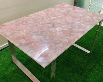 以下が含まれることがあります： 銀色の金属フレームにピンクのクォーツの天板が付いたテーブル。天板は、研磨され、パターン状に配置された天然のクォーツクリスタルで作られています。テーブルは緑の人工芝の上に置かれています。