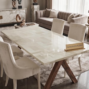 Puede incluir: Una mesa de comedor rectangular con una superficie de piedra blanca pulida con patrón de espiga y una base de madera marrón oscuro en forma de X. Un pequeño letrero rectangular dorado con el texto GemsAndArtGallery descansa sobre la mesa.