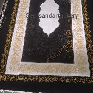 Puede incluir: Una tela negra con un dise&ntilde;o bordado en blanco y dorado. El dise&ntilde;o presenta un motivo floral central con fondo blanco y contornos dorados. La tela est&aacute; estirada en un marco de madera.