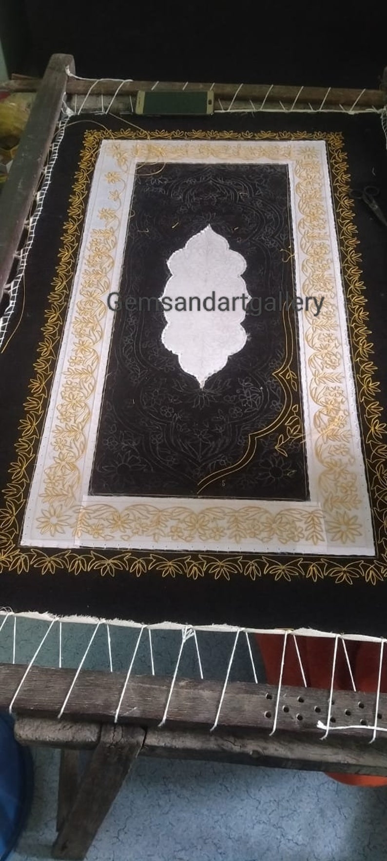 Puede incluir: Una tela negra con un dise&ntilde;o bordado en blanco y dorado. El dise&ntilde;o presenta un motivo floral central con fondo blanco y contornos dorados. La tela est&aacute; estirada en un marco de madera.
