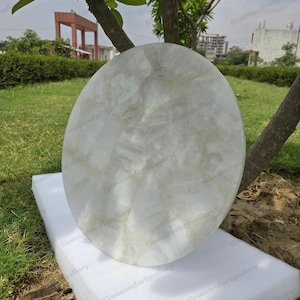 May include: A large, round, white stone table top with subtle gray veining. The table top is resting on a white foam surface, with a green grassy background and a tree in the foreground. The table top is approximately 30 inches in diameter.
