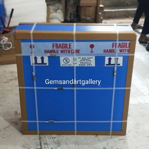 Op de afbeelding: Een blauwe kartonnen doos met bruine tape en witte banden. De doos is gelabeld met "FRAGILE HANDLE WITH CARE" en "FI AGILI HANDL WITH". De doos heeft ook een sticker met de tekst "DO NOT DROP HANDLE WITH CARE FRAGILE KEEP DRY".