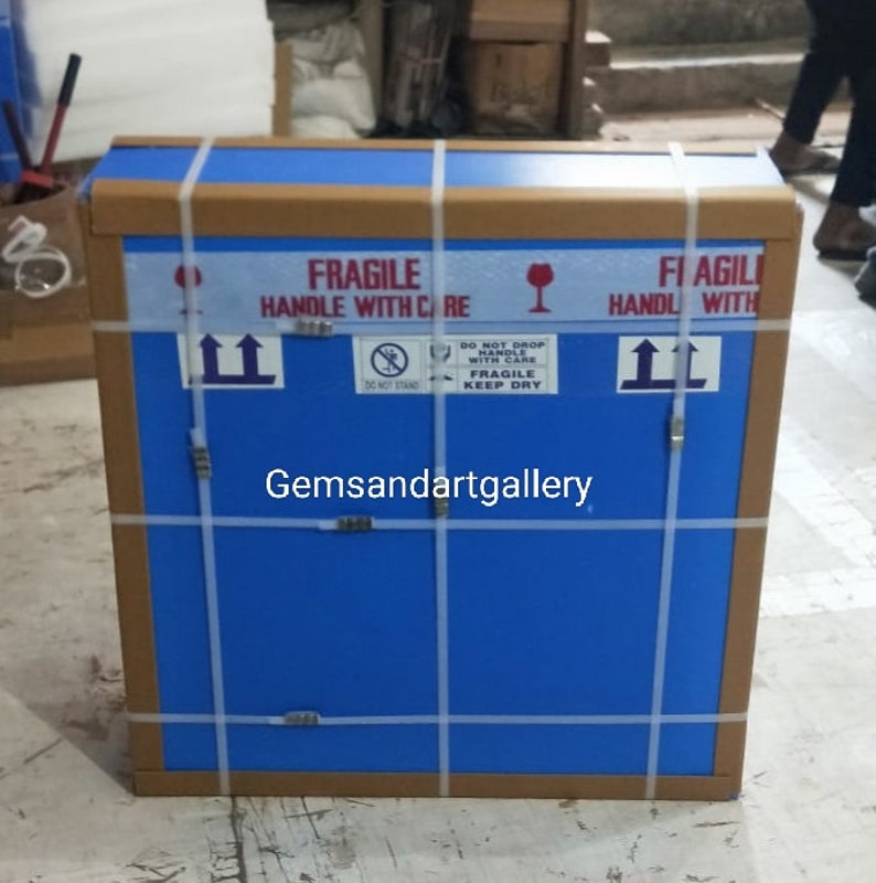 Op de afbeelding: Een blauwe kartonnen doos met bruine tape en witte banden. De doos is gelabeld met "FRAGILE HANDLE WITH CARE" en "FI AGILI HANDL WITH". De doos heeft ook een sticker met de tekst "DO NOT DROP HANDLE WITH CARE FRAGILE KEEP DRY".