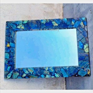 May include: A rectangular mirror with a frame made of blue and green labradorite gemstone pieces. The mirror has a light blue reflection.