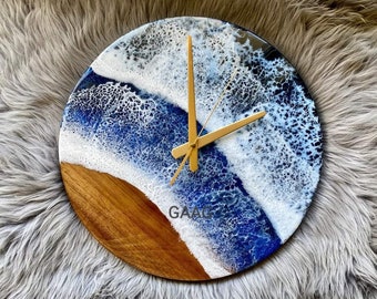 Può includere: Un orologio rotondo in legno con un quadrante in resina blu e bianco che rappresenta le onde dell'oceano. Le lancette dell'orologio sono dorate.