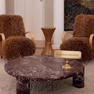 Könnte beinhalten: Zwei braune Schaffell-Sessel mit Armlehnen aus hellem Holz, ein runder Couchtisch aus dunkelviolettem Marmor mit zylindrischen Beinen und ein kleiner Beistelltisch aus Holz. Ein Messingschild zeigt den Text GemsAndArtGallery.