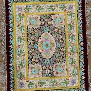 Puede incluir: Una pequeña alfombra cuadrada de terciopelo burdeos con un diseño floral dorado y blanco. La alfombra está bordada con cuentas y lentejuelas de colores. La alfombra tiene un borde de flecos.