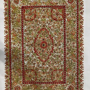Puede incluir: Un cuadrado de tela blanca con intrincados bordados en hilo dorado, rojo y verde. El dise&ntilde;o presenta una forma de diamante central con motivos florales y un borde de patrones repetidos.