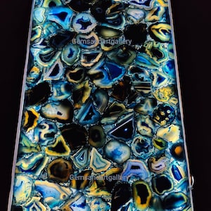 May include: A close-up of a blue and gold agate slab with a polished surface. The slab is made up of many small pieces of agate that have been arranged in a mosaic pattern. The agate is backlit, which makes the colors appear more vibrant. The text "Gemsandartgallery" is visible in the image.