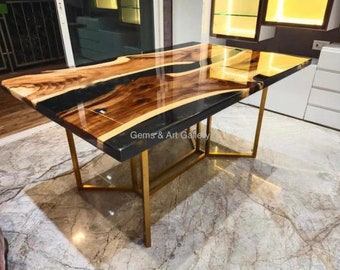May include: A rectangular dining table featuring a live edge wood slab with a glossy finish and a black resin river inlay. The table is supported by a gold-coloured metal frame with angular legs. The text Gems & Art Gallery is visible on the surface.