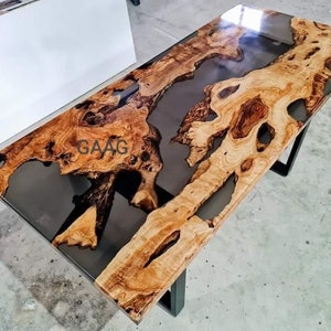 May include: A large wooden table with a black epoxy resin river flowing through the center. The table has a natural wood finish and is supported by black metal legs. The table is approximately 8 feet long and 3 feet wide.