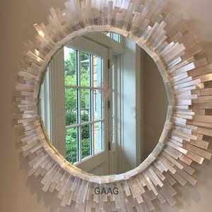 May include: A round mirror with a white selenite crystal frame. The mirror is reflecting a window with a view of trees outside.