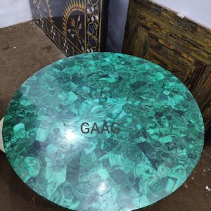 May include: A round table top made of malachite stone. The surface is a mosaic of green and dark green pieces, creating a unique pattern. The text "GAAC" is visible on the surface. The table top is a decorative element.