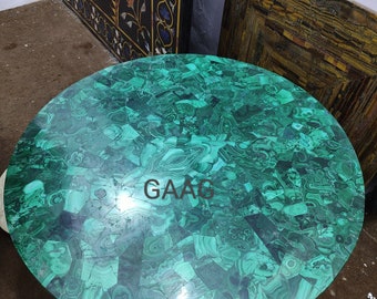 May include: A round table top made of malachite stone. The surface is a mosaic of green and dark green pieces, creating a unique pattern. The text "GAAC" is visible on the surface. The table top is a decorative element.