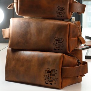 May include: Three brown leather zippered pouches stacked on top of each other. Each pouch has a black leather strap and a zipper closure. The pouches are decorated with the text "Home is where you are" in a black script font.