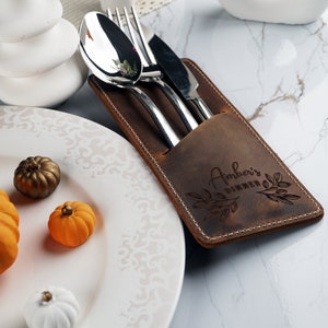 May include: A brown leather cutlery holder with the text "Amber's Dinner" engraved on it. The holder has a pocket for a spoon, fork, and knife.