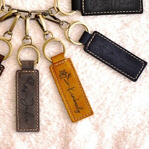 May include: A collection of personalized leather keychains in various colors, each featuring a name and floral design. Keychains have brass-colored hardware and white stitching. Colors include brown, red, dark brown, gray, yellow, black, and navy.