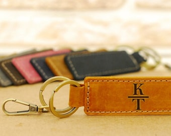 May include: Leather keychains in various colors, including brown, black, and pink. The focus is on a tan leather keychain with the initials "KT" engraved. The keychain has a metal clasp and ring.
