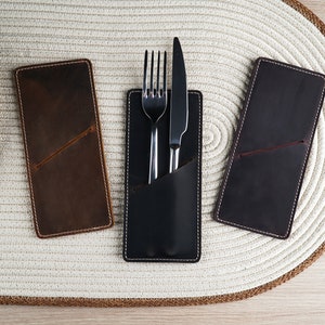 May include: Three leather cutlery holders, one brown, one black, and one dark brown. The black holder has a fork and knife inside.