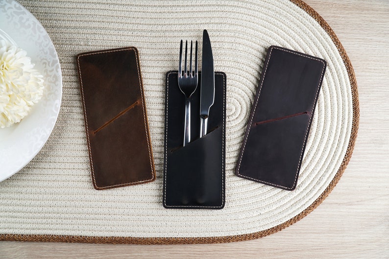 May include: Three leather cutlery holders, one brown, one black, and one dark brown. The black holder has a fork and knife inside.