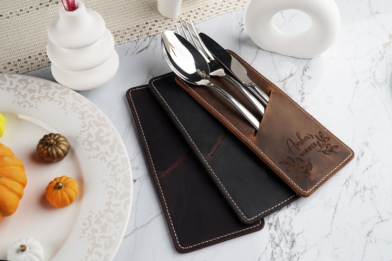 May include: A brown leather cutlery holder with a personalized inscription "Amber's Kitchen" and a decorative leaf design. The holder contains a fork, knife, and spoon.