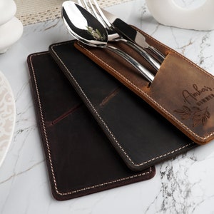 May include: Two leather cutlery holders, one brown and one black, with a set of silverware inside the brown holder. The brown holder has the text "Amber's Dinner Club" engraved on it.