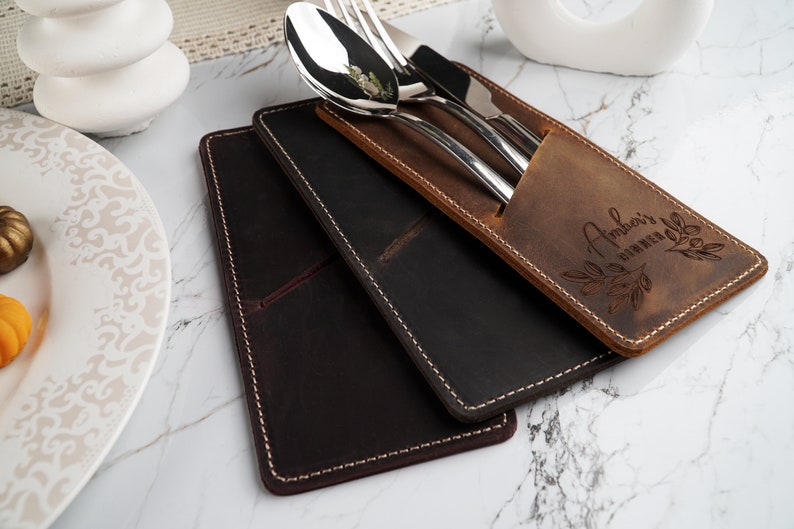 May include: Two leather cutlery holders, one brown and one black, with a set of silverware inside the brown holder. The brown holder has the text "Amber's Dinner Club" engraved on it.